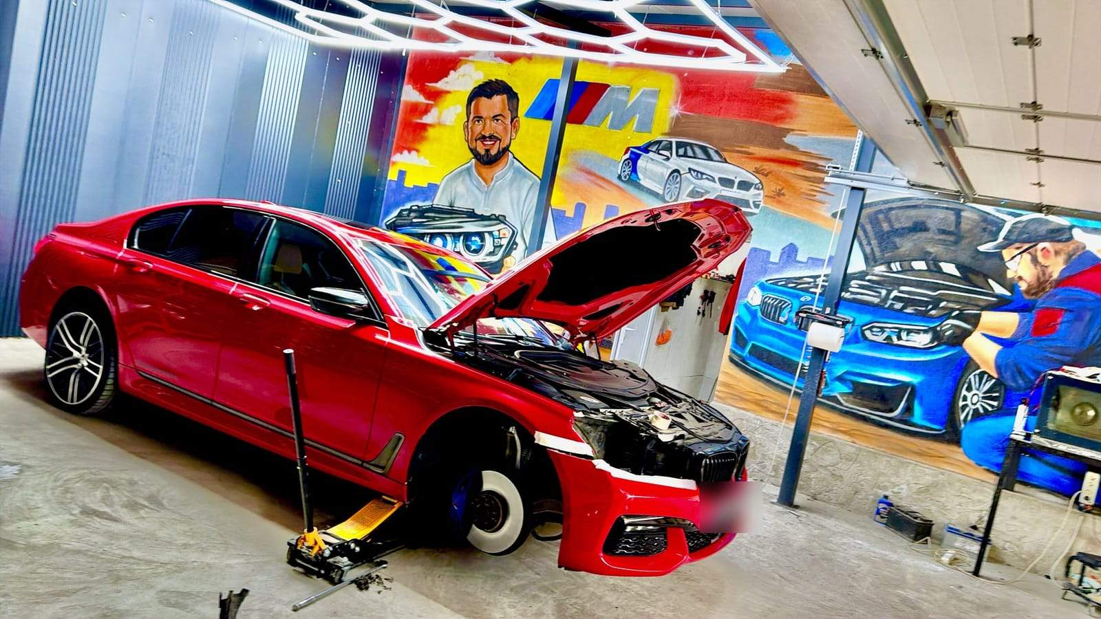 Atelierul Low Garage: un BMW roșu cu capota ridicată sub plafonul de lumini hexagonale, în fața muralului cu mașini BMW